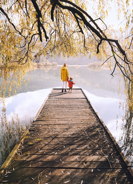 Femme et enfant sur un pont en bois près du lac entouré de branches de saule en automne