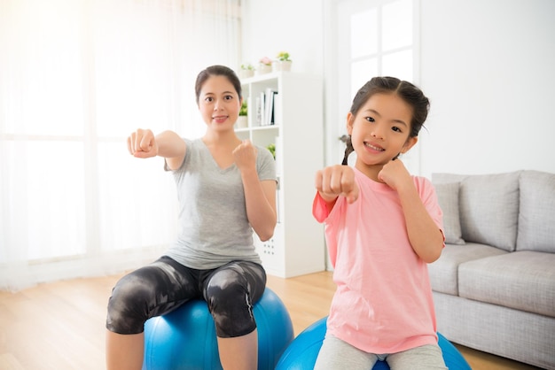 la femme et les enfants font un geste des mains comme de la boxe et frappent l'air en s'asseyant sur l'équilibre et la force d'entraînement du ballon de fitness dans le cours de yoga professionnel au centre de gym.