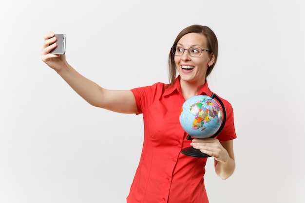 Femme enseignante d'affaires souriante en chemise rouge tenant un téléphone portable et faisant une prise de selfie avec un globe isolé sur fond blanc. Enseignement de l'éducation dans le concept d'université de lycée. Espace de copie.