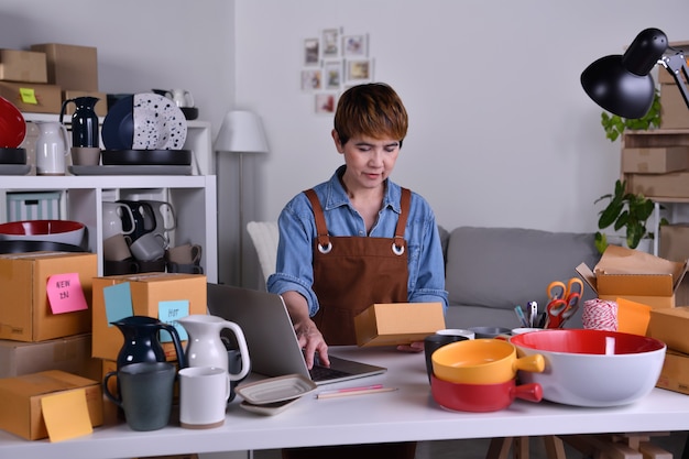 Femme entrepreneur asiatique mature, propriétaire d'entreprise travaillant sur un ordinateur portable vérifiant l'adresse et les détails de livraison avant d'expédier le produit. Concept de travail d'entreprise de vente en ligne à la maison