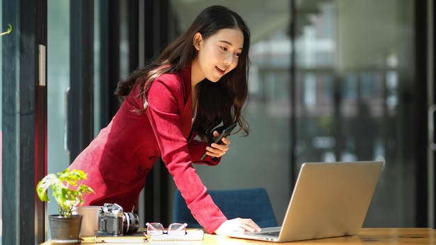 Une femme entrepreneur est satisfaite des performances de vente et des bénéfices de son entreprise alors qu'elle s'appuie contre son bureau et regarde le rapport sur son ordinateur portable.