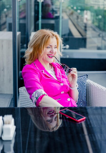 Femme entrepreneur prospère dans une veste magenta posant sur la terrasse d'un café dans un centre d'affaires