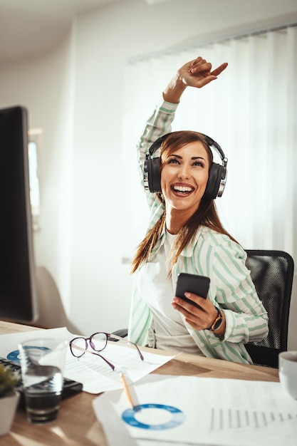 Une femme entrepreneur prospère avec des écouteurs sur la tête écoute de la musique sur un smartphone au bureau.