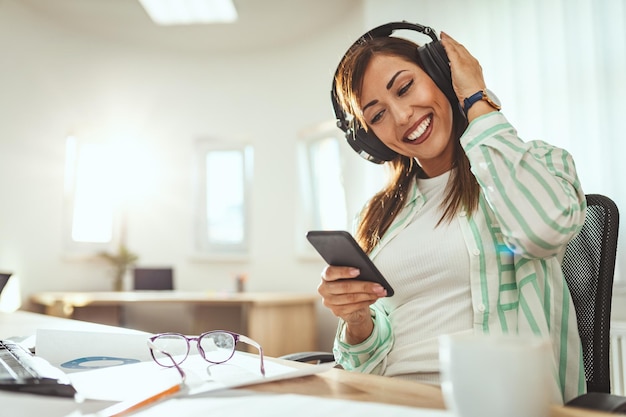 Une femme entrepreneur prospère, avec des écouteurs sur la tête, recherche de la musique sur un smartphone au bureau.