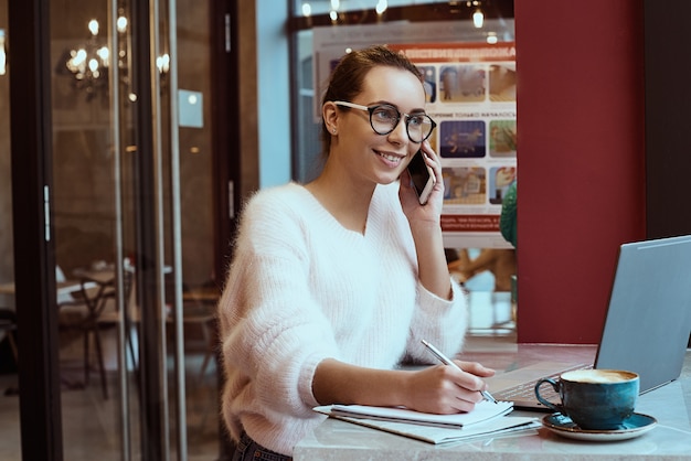 Femme entrepreneur travaillant dans un café et parlant sur téléphone mobile avec des écouteurs