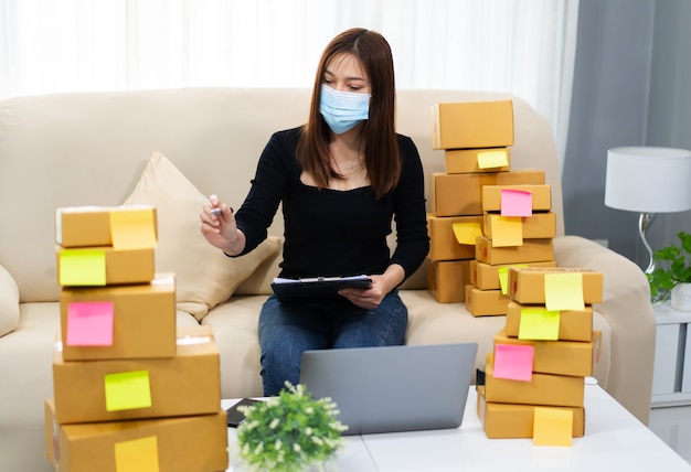 Femme entrepreneur travaillant avec un ordinateur portable et vérifiant pour préparer la boîte à colis pour la livraison au client au bureau à domicile, il porte un masque facial pour protéger la pandémie de coronavirus