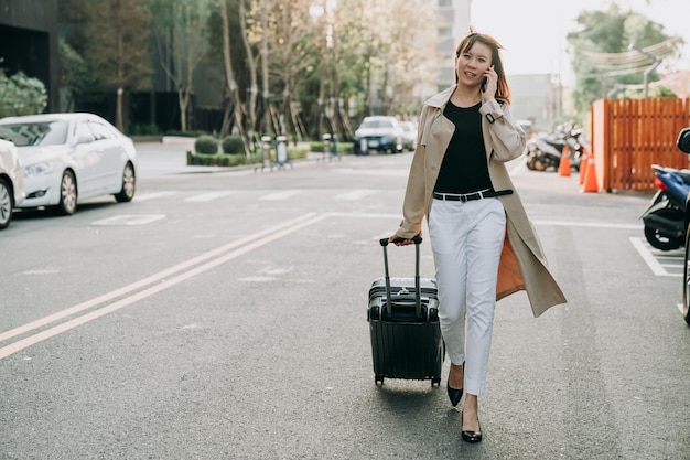 Une femme entrepreneure asiatique élégante de pleine longueur marchant dans la rue de la ville a une conversation sur un smartphone tout en partant pour un voyage d'affaires avec une valise par une journée ensoleillée.