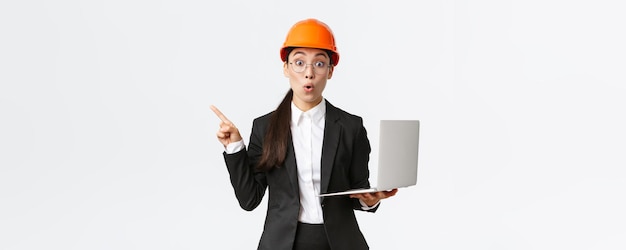 Femme entrepreneure asiatique impressionnée à l'usine portant un casque de sécurité et un costume d'affaires pointant le doigt vers la gauche et tenant un ordinateur portable montrant un diagramme à l'entreprise fond blanc