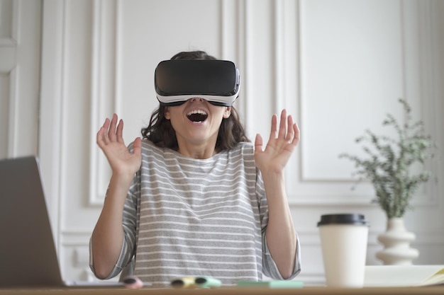 Une femme entrepreneure enthousiaste teste la technologie de réalité virtuelle portant des lunettes vr assise au bureau à domicile