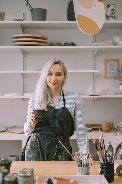 Femme entrepreneure portant un tablier travaillant dans un atelier de poterie et un magasin de céramique Femme artisan et propriétaire d'une petite entreprise d'artisanat parmi des objets de poterie en céramique faits à la main dans l'espace de travail d'un studio d'art