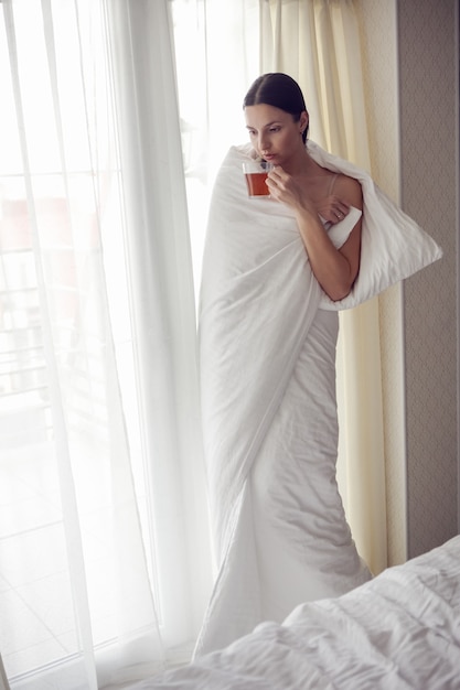 Photo une femme enveloppée dans une couverture blanche se tient à côté d'une grande fenêtre , des rideaux boit du thé dans une tasse se réchauffant du froid après un bain