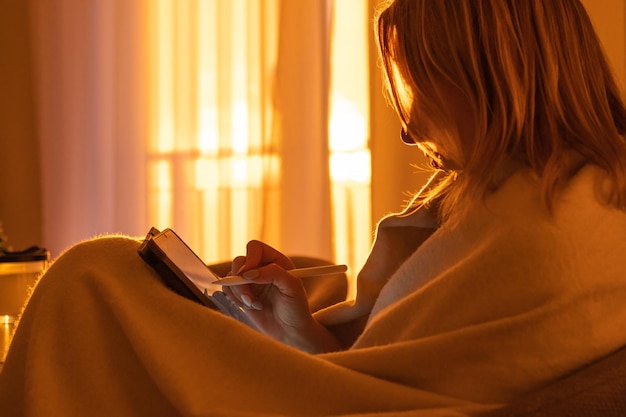 Femme enveloppée dans la couverture et utilise une tablette assise dans le salon à la lumière du coucher du soleil