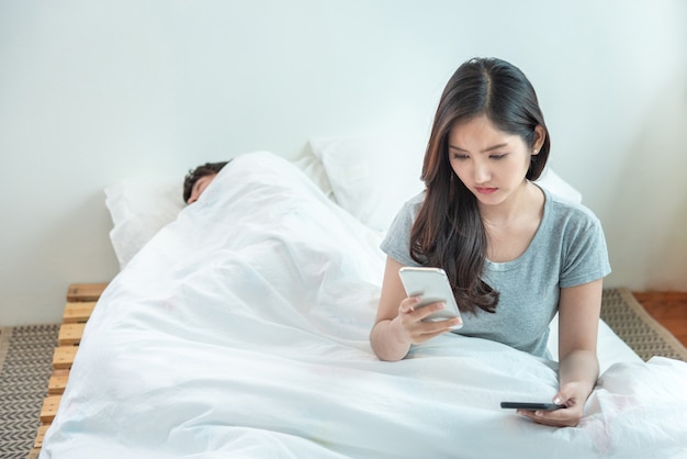 Femme espionnant le téléphone de son mari pendant que l'homme dort dans son lit à la maison.Contrôle et suspension de la jeune fille asiatique sur le téléphone de son petit ami pendant qu'il fait la sieste sur le lit.