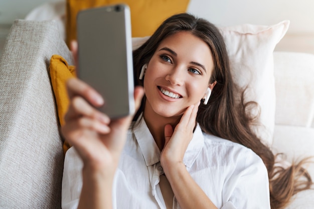 Photo la femme est allongée sur un canapé, se détend, utilise un smartphone pour des messages texte, échange des photos, regarde des vidéos.