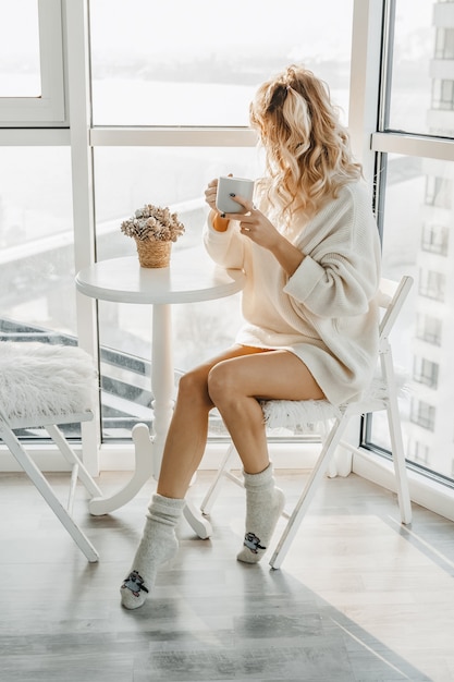 La femme est assise et boit du café dans un intérieur clair.