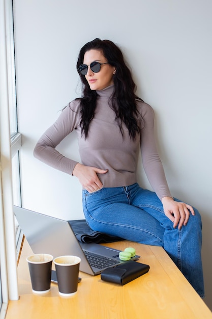 Une femme est assise sur un bureau devant une fenêtre avec un ordinateur portable et une tasse de café.