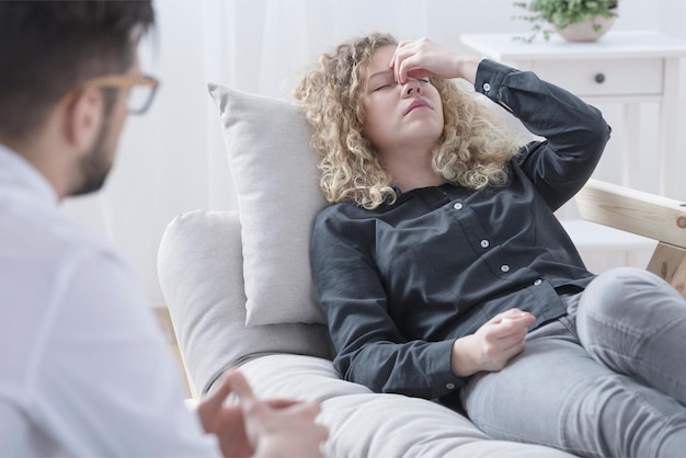 Une femme est assise sur un canapé avec sa main sur son front et sa tête est examinée par un médecin.