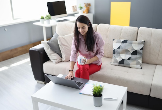 Une femme est assise sur le canapé, tenant une tasse d'écrevisses et, avec son autre main, tapant sur un ordinateur portable