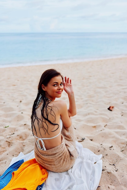 Une femme est assise avec le dos à la caméra après avoir nagé dans l'océan en maillot de bain sur une serviette sur le sable et regarde la mer et les nuages vacances d'été à la plage