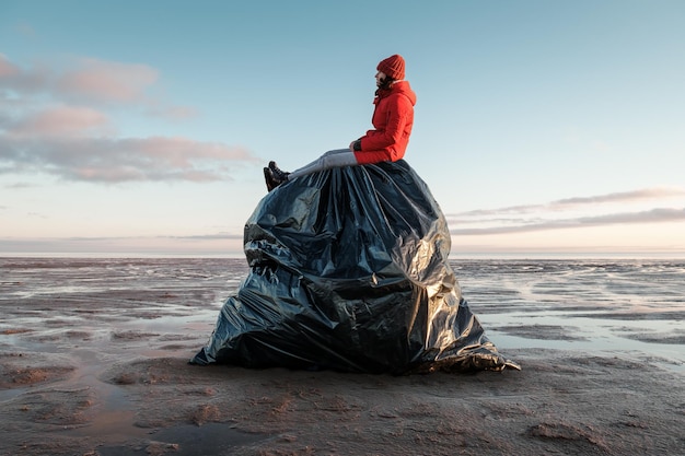 Une femme est assise sur un énorme sac poubelle noir sur le rivage