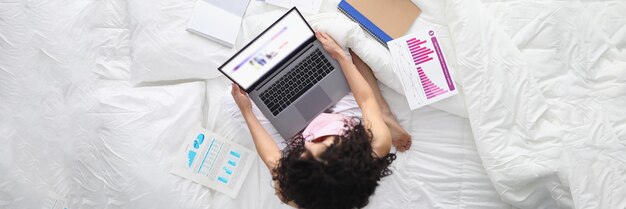 Une femme est assise sur le lit avec un ordinateur portable et des documents