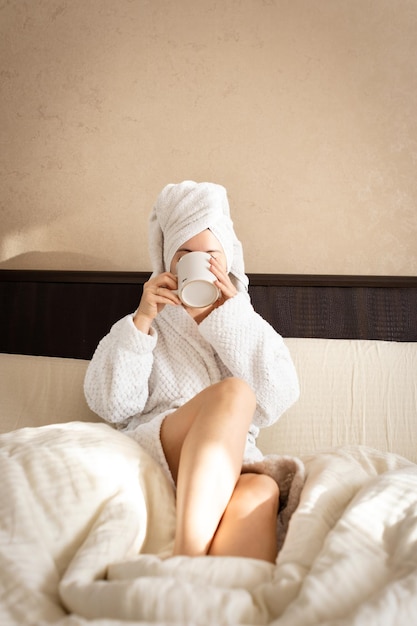 Une femme est assise sur un lit vêtue d'une robe de chambre blanche avec une serviette couvrant sa tête et boit du thé