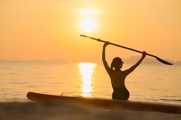 La femme est assise sur la plage avec le sup board à côté d'elle au coucher du soleil