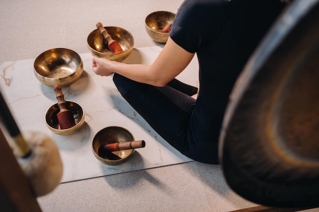 Une femme est assise en position du lotus à côté de bols tibétains assis sur un tapis de yoga sur fond de gong