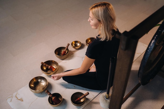 Une femme est assise en position du lotus à côté de bols tibétains assis sur un tapis de yoga sur fond de gong