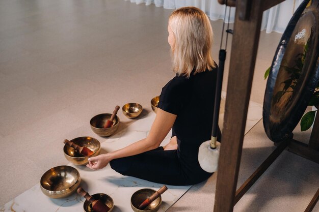 Une femme est assise en position du lotus à côté de bols tibétains assis sur un tapis de yoga sur fond de gong