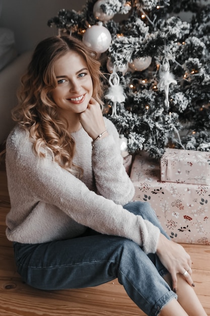 La femme est assise près de l'arbre de Noël et présente. Elle sourit.