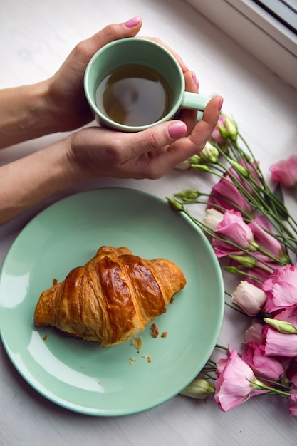 Femme est assise près de la fenêtre avec des fleurs roses et un croissant sur une plaque verte