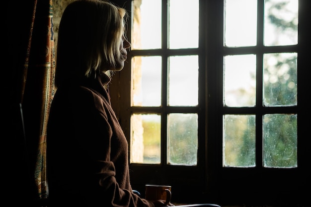 Une femme est assise près de la fenêtre avec une tasse de café Concept de tristesse de solitude Réflexions sur la crise de la quarantaine