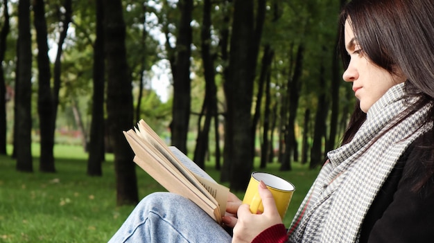 Une femme est assise sous un arbre et lit son livre préféré en buvant du café ou du thé dans une tasse jaune dans un parc de la ville sur l'herbe verte par une agréable journée ensoleillée. Concept de vacances, d'éducation et d'étude.