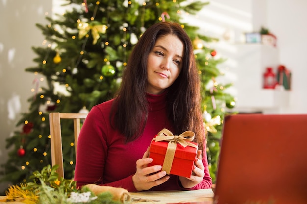 La femme est assise à la table et déballe le cadeau de Noël Sapin de Noël en arrière-plan célébration en ligne