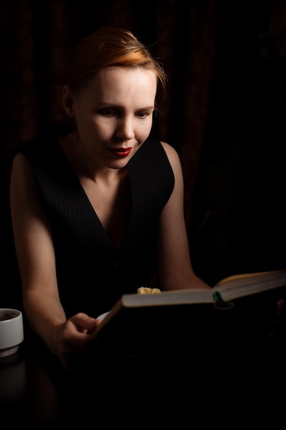 Une femme est assise à une table avec un livre et du thé et des gâteaux dans le noir Une femme de quarante ans se détend seule