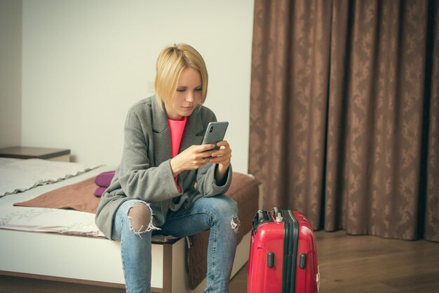 Une femme est assise avec un téléphone sur le lit près de la valise