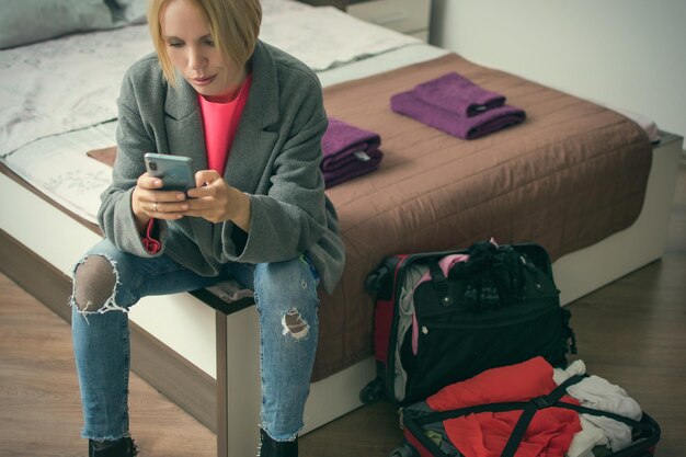 Une femme est assise avec un téléphone sur le lit près de la valise