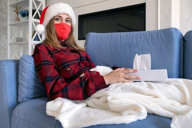 La femme est malade à Noël. Jeune femme blonde dans une chemise rouge et une casquette de Noël portant un masque médical de protection se trouve sur le canapé avec des serviettes