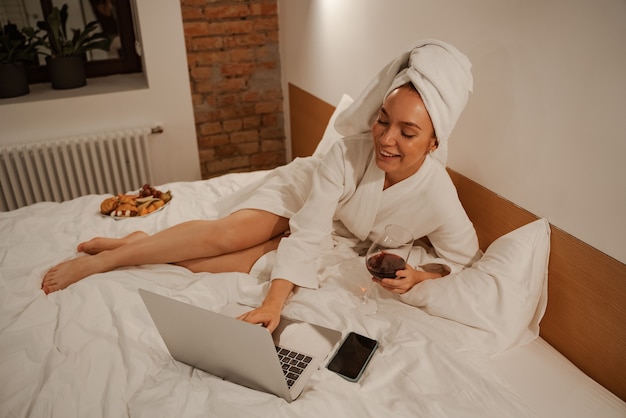 La femme est vêtue d'une robe et d'une serviette blanche sur la tête. Elle s'allonge sur le lit et surfe sur Internet avec un verre de vin.