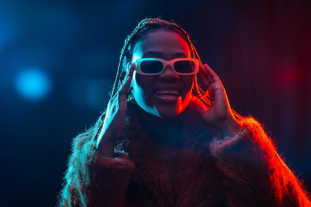 Femme ethnique noire avec des tresses avec des lumières led bleues et rouges portant un manteau de fourrure souriant