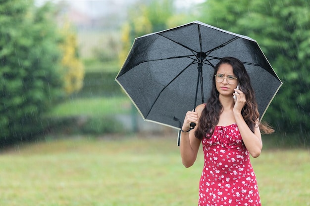 Femme ethnique avec parapluie ayant une dispute téléphonique dans le parc le jour de la pluie