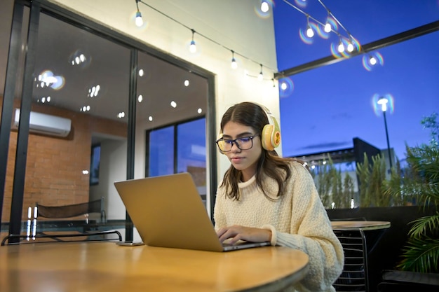 Une femme ethnique travaille en ligne depuis votre ordinateur portable à l'extérieur dans un café au crépuscule