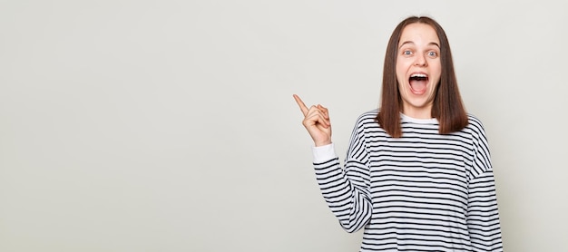 Femme étonnée aux cheveux bruns portant une chemise rayée criant d'excitation pointant vers l'espace de copie pour la zone de publicité de promotion debout isolée sur fond gris