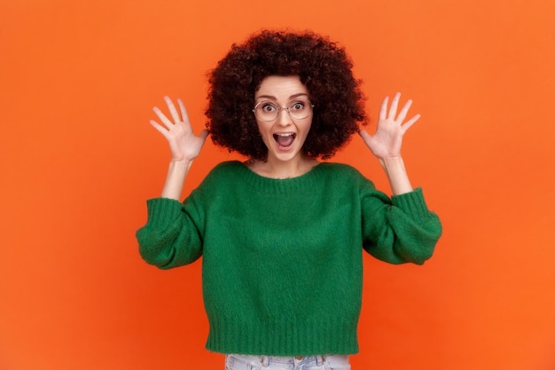 Femme étonnée avec une coiffure Afro portant un pull vert de style décontracté et des lunettes debout avec les bras levés, hurlant d'excitation. Studio intérieur tourné isolé sur fond orange.