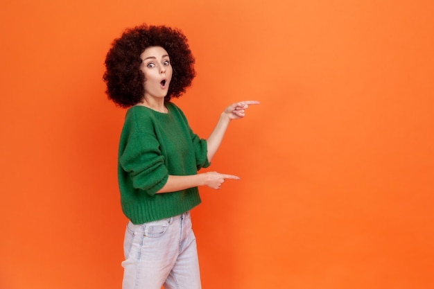 Femme étonnée avec une coiffure Afro en pull vert pointant de côté avec le doigt, ayant surpris l'expression du visage, garde la bouche ouverte, copie l'espace. Studio intérieur tourné isolé sur fond orange.