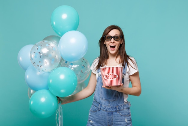 Femme étonnée dans des lunettes 3d regardant un film, tenez un seau de pop-corn célébrant avec des ballons à air colorés isolés sur fond bleu turquoise. Fête d'anniversaire, émotions dans le concept de cinéma.