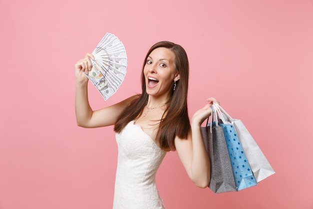 Une femme étonnée en robe blanche tient un paquet de dollars, de l'argent en espèces, des sacs multicolores avec des achats après le shopping