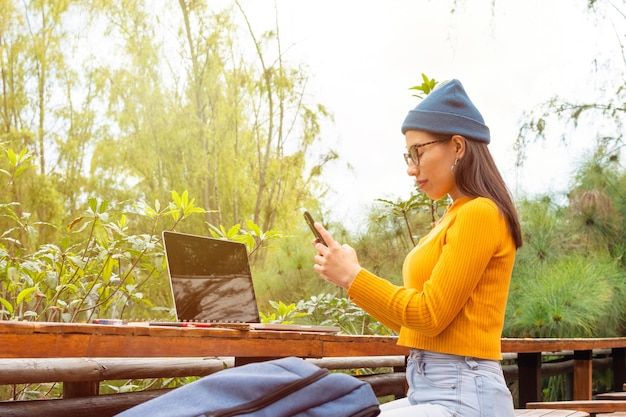 Femme étudiante latina tapant sur son téléphone portable devant son ordinateur portable à l'extérieur pendant une journée ensoleillée