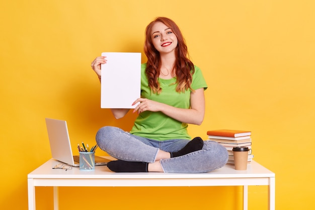 Femme étudiante souriante assise sur la table et tenant une chemise en papier vierge dans les mains